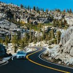 A car drives through a winding road in a mountainous landscape, surrounded by trees and rocks under a clear summer sky. Road Trip