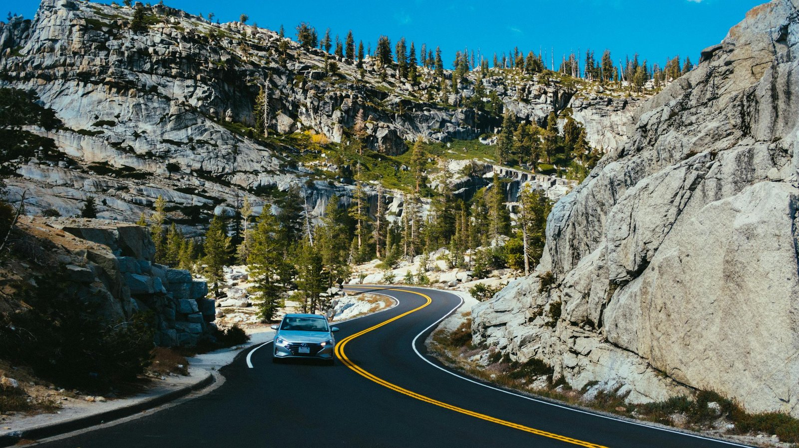 A car drives through a winding road in a mountainous landscape, surrounded by trees and rocks under a clear summer sky. Road Trip