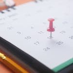 A close-up of a calendar page with a pink push pin, pencil, and binder clip on an orange background. Travel Dates