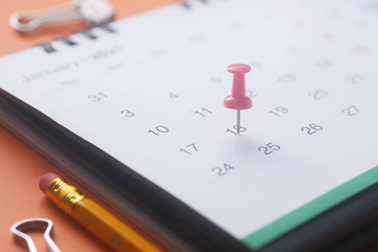 A close-up of a calendar page with a pink push pin, pencil, and binder clip on an orange background. Travel Dates