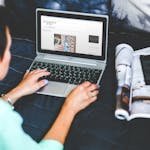 A woman on a bed browsing and blogging with a laptop and a magazine nearby. - travel research
