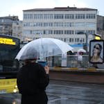 A woman waits by a bus stop in Istanbul under a clear umbrella on a rainy day.