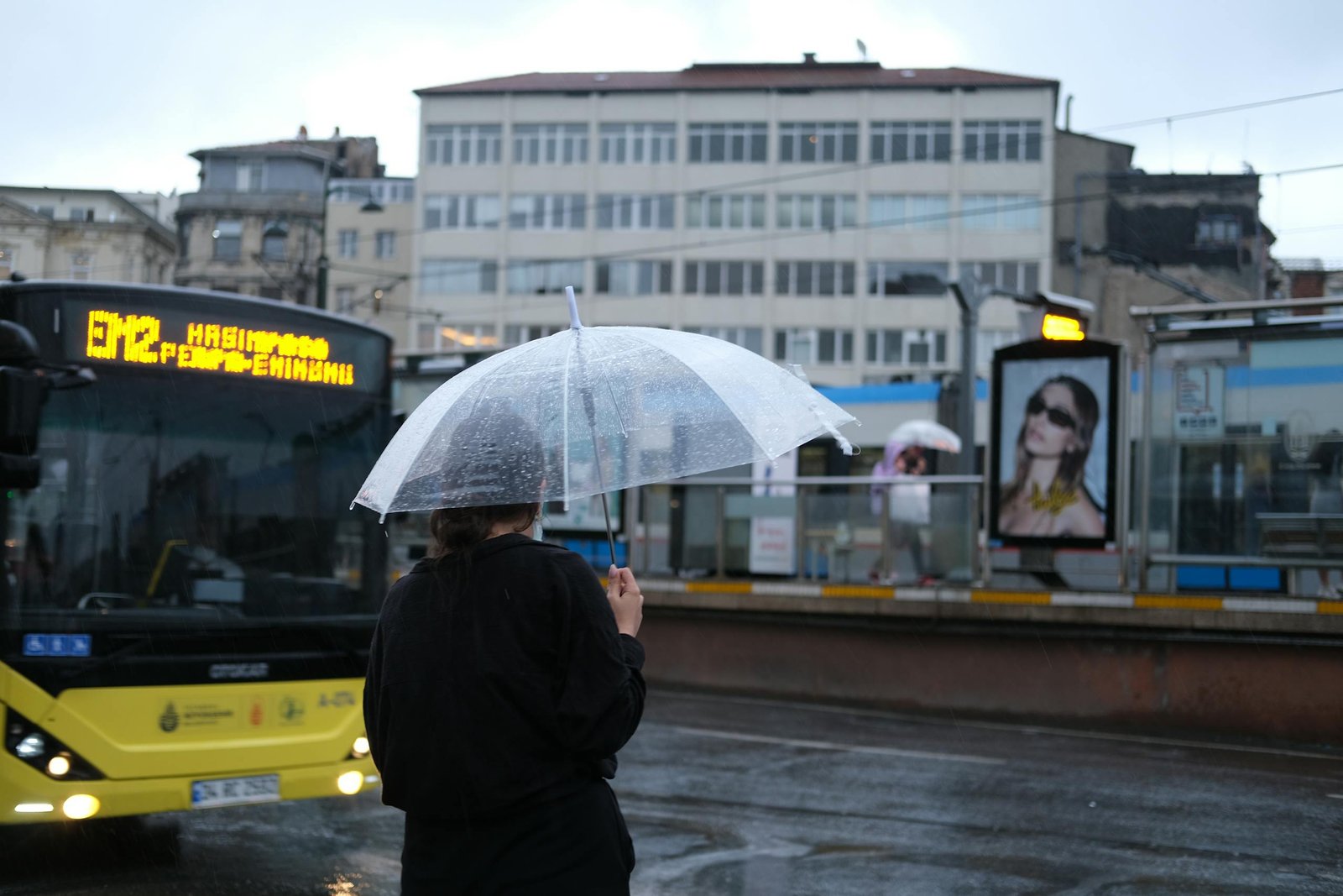 A woman waits by a bus stop in Istanbul under a clear umbrella on a rainy day.