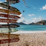 Colorful signpost on beach showing distances to various cities, sunny day.