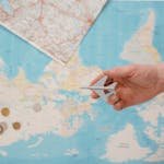 Hand holding a toy airplane over a world map, surrounded by coins, camera, and passport.