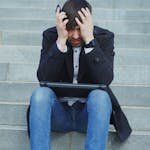 Man sitting on stairs with laptop, looking stressed and frustrated.