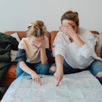 Mother and daughter covering eyes while pointing at map on couch, planning a trip.