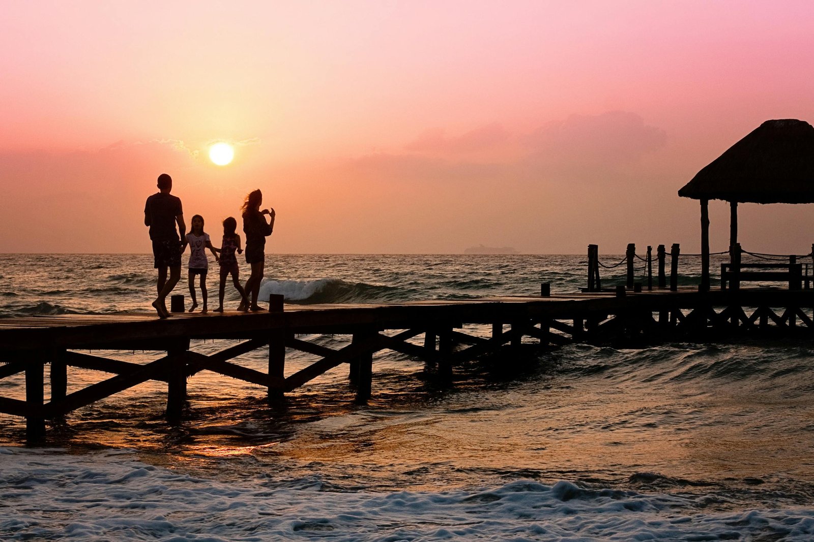 Silhouetted family enjoys a stroll on the beach pier at a vibrant sunset over the ocean waves. - Family Vacation