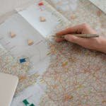 Hand holding pen over Europe map, planning travel route with markers.