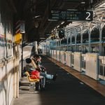 Commuters seated on a platform at a busy Tokyo train station under warm daylight.