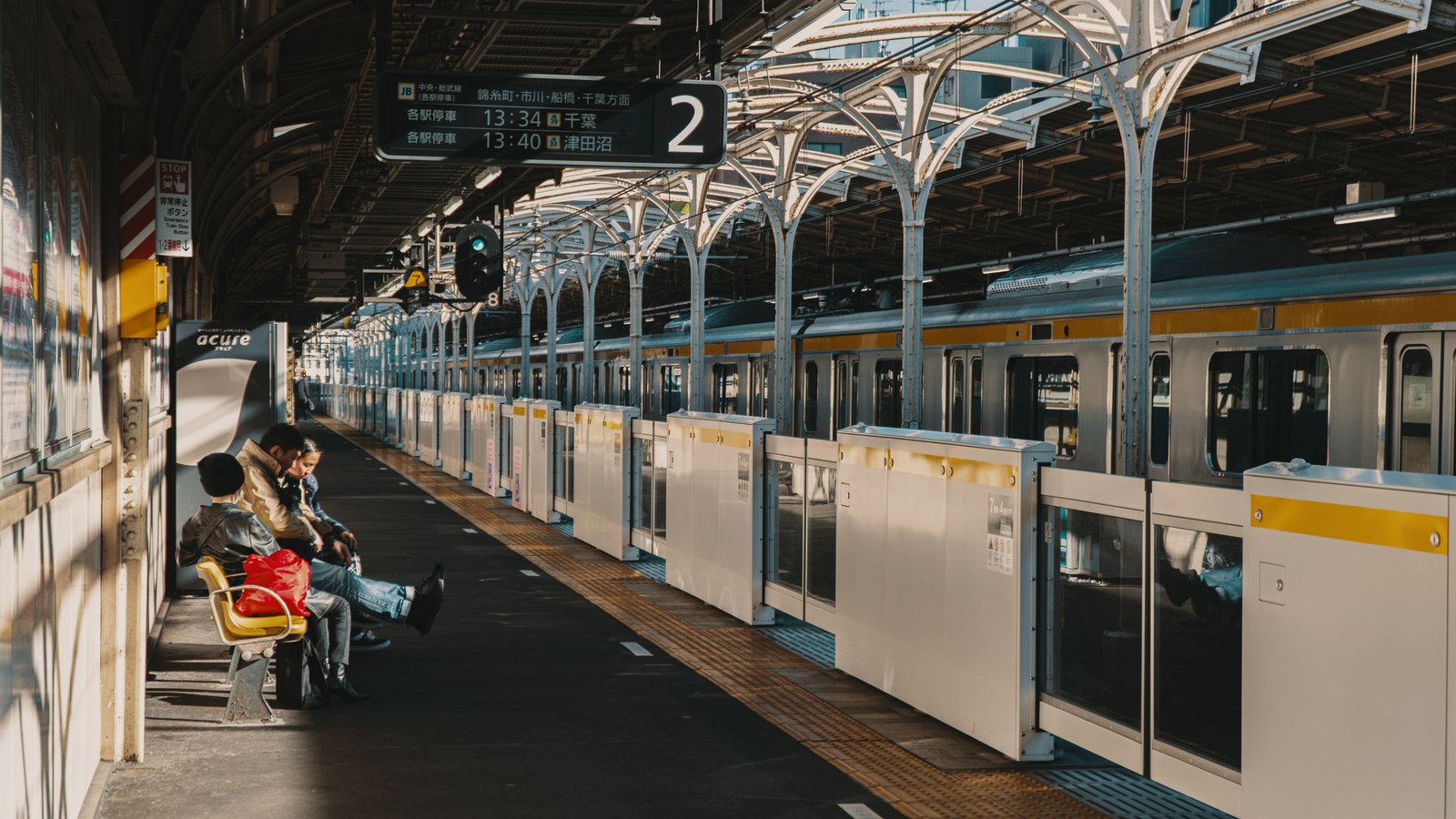 Commuters seated on a platform at a busy Tokyo train station under warm daylight.