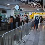 Travelers passing through airport security with staff and metal railings visible.