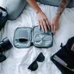 Woman with tattoos packs electronics, camera, and sunglasses in a suitcase on a bed.